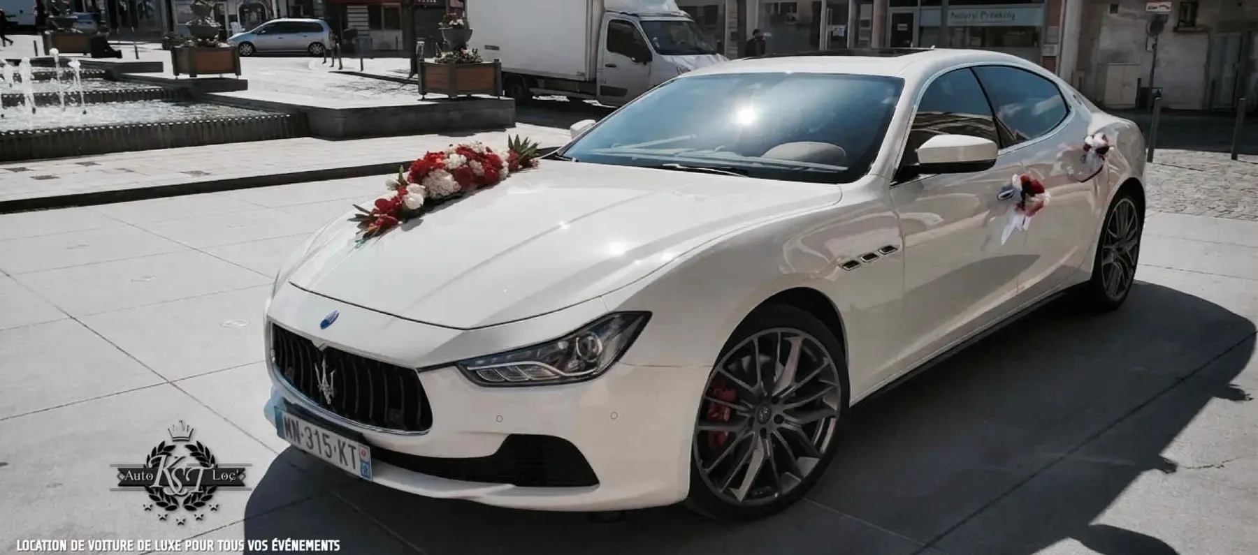 Maserati Ghibli blanche décorée de fleurs rouges et blanches, vue avant trois-quarts sur une place
